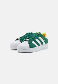 Green and white sneakers with a textured suede upper, rubber shell toe, three white stripes, and gold accent on the lacing.