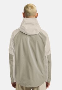 Persona con capelli biondi corti che indossa una giacca con cappuccio beige bicolore, vista di spalle su uno sfondo chiaro e semplice.