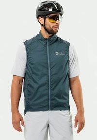 Σκοτεινό τυρκουάζ, αμάνικο γιλέκο ποδηλασίας με φερμουάρ μπροστά, δύο πλαϊνές τσέπες και λεπτομέρεια λογότυπου. Φορεμένο πάνω από ένα ελαφρύ γκρι μπλουζάκι με κοντά μανίκια.