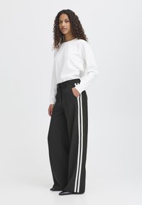 Pantaloni neri a gamba larga con strisce bianche ai lati, abbinati a un top bianco a maniche lunghe. Il tessuto sembra liscio; i pantaloni presentano una vita alta e tasche.