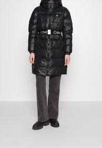 Svart puffjacka med huva, belagd midja, dragkedja och Calvin Klein-logotyp på bröstet; slät, glänsande yta.