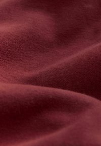 Bordeaux stof met een gladde textuur, met een fijn breipatroon. Het materiaal lijkt iets gekreukt, wat de zachte val benadrukt.