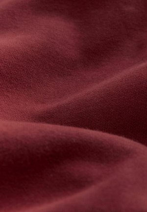 Bordeaux stof met een gladde textuur, met een fijn breipatroon. Het materiaal lijkt iets gekreukt, wat de zachte val benadrukt.