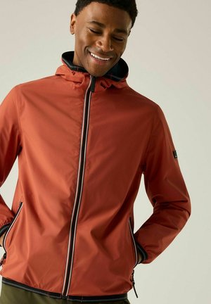 Mann lächelt und schaut nach unten, trägt eine rostfarbene Zip-Hoodie-Jacke mit Händen in den Taschen vor einfarbigem Hintergrund.