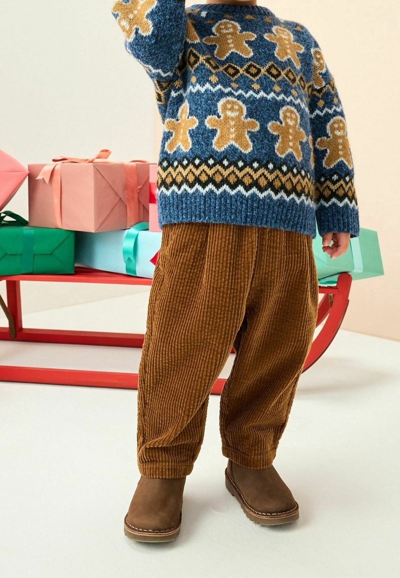 Enfant portant un pull bleu avec des bonhommes en pain d'épice, pantalon en velours marron et bottes marron, debout près d'un traîneau rouge avec des cadeaux emballés.