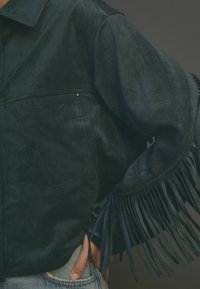Dunkelgrüne Wildlederjacke mit einem Kragen, zwei Vorderas Taschen und Fransenverzierungen an den Ärmeln. Weiche Textur mit matter Oberfläche.