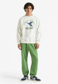 Felpa bianca con stampa grafica verde e blu; pantaloni della tuta verdi; abbinati a sneaker blu scuro; vestibilità morbida e casual.