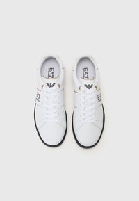 Scarpe sportive bianche con una texture simile alla pelle liscia, dotate di occhielli dorati, suole nere e logo sui lati e sulle solette.