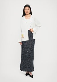 White button-up jacket with a Tweety Bird print pocket, paired with a long black floral-patterned skirt and black pointed-toe shoes.