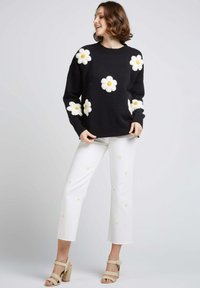 Un pull noir avec des marguerites blanches et des cœurs jaunes, associé à un pantalon blanc cropped avec des broderies florales. Des sandales à talons hauts beiges.