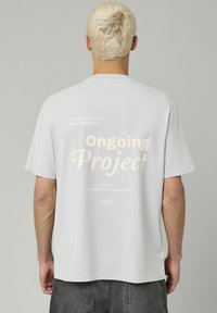 T-shirt en coton gris avec un grand texte graphique crème "Projet en cours" au dos, accompagné d'autres phrases en petits caractères au-dessus.