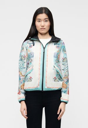 Femme portant une veste blanche zippée avec des motifs colorés floraux et animaliers, capuche noire et pantalon noir, debout devant un fond uni.