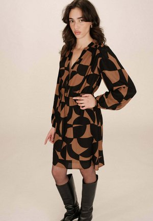 Robe à manches longues avec un motif géométrique noir et marron, présentant un col en V, des boutons sur le devant et une taille élastique. Bottes noires montantes jusqu'au genou.