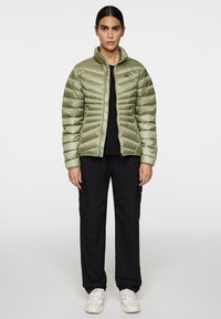 Vaaleanoliivinvihreä puffatakki korkealla kauluksella, vetoketjukiinnityksellä ja tikkikuvioinnilla, yhdistettynä mustiin cargo-housuihin ja valkoisiin tennareihin.