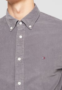 Camisa de pana gris con cuello abotonado, que cuenta con botones blancos y un pequeño logo bordado en el lado izquierdo del pecho.
