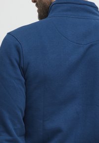 Blå hettegenser med høy krage, laget av et teksturert, mykt stoff med sømdetalj på baksiden. Enkel, ensfarget design.