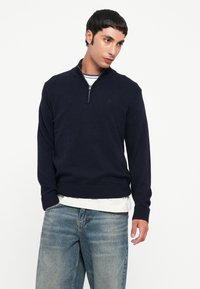 Mann, der einen marineblauen Zip-Pullover über einem gestreiften Hemd und verblassten blauen Jeans trägt, vor einem schlichten weißen Hintergrund steht.