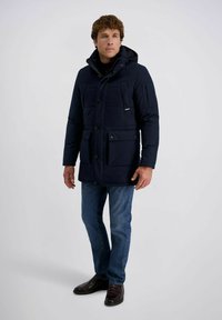 Marineblauwe gewatteerde jas met een capuchon, meerdere zakken en knoopsluitingen. Draagt over een zwarte coltrui met blauwe spijkerbroek en donkere schoenen.