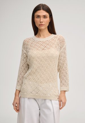 Vrouw draagt een beige gehaakte trui met driekwart mouwen en witte losse broek, staand tegen een effen lichte achtergrond.