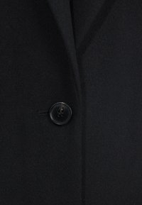 Abrigo de lana negro con una textura suave, solapas marcadas y un gran botón negro en el frente, que muestra un diseño a medida.