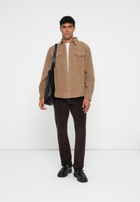 Bruine corduroy shirtjack met voorzakken, gecombineerd met donkerbruine broek en zwarte schoenen. Een zwarte schoudertas wordt aan de linkerzijde gedragen.