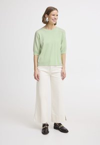 Maglietta a maniche corte di colore verde chiaro abbinata a pantaloni larghi di colore bianco sporco e mocassini neri, con dettagli in hardware dorato.