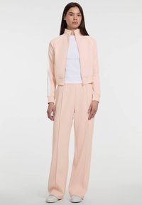 Lichtroze trainingspak met een cropped ritsjack met witte details en bijpassende losse broek met een gladde textuur. Witte schoenen.