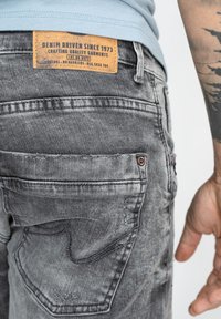 Grå denimjeans har en texturerad yta, bakfickdesign med subtila stygn och en synlig brun läderetikett på midjebandet.