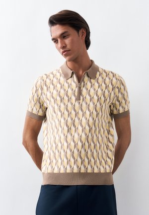 Άνδρας όρθιος με τα χέρια πίσω από την πλάτη, φορά κοντομάνικο πόλο πουκάμισο με γεωμετρικό μοτίβο σε μπεζ, κίτρινο και καφέ χρώμα, και σκούρο παντελόνι.