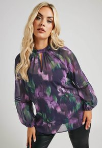 Transparent lila blus med blommönster som har en nyckelhålsöppning, långa ärmar med elastiska manschetter och en flödande passform. Mönster med gröna och lavendelfärgade blommor.