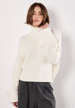 Crèmekleurige ribgebreide turtleneck trui met oversized mouwen, gemaakt van een textuurrijke, ademende stof en een ontspannen pasvorm. Gecombineerd met grijs gestreepte broek.