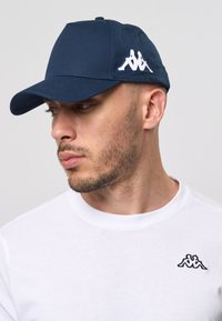 Marineblå baseballcap med en strukturert design, med en hvit brodert logo på siden. Laget av slankt, slitesterkt stoff.