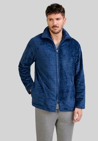 Chaqueta de cremallera azul marino hecha de tejido de felpa texturizada con un patrón de cuadros, que cuenta con un cuello alto y dos bolsillos laterales.