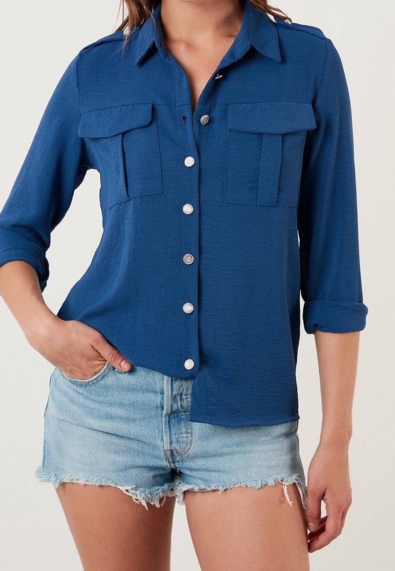 Femme portant une chemise bleue boutonnée avec manches retroussées et deux poches poitrine, associée à un short en jean bleu clair effiloché.