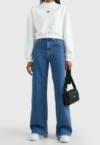 Sudadera blanca corta con mangas largas y un pequeño logo, combinada con jeans azules de pierna ancha y cintura alta, y un bolso cruzado negro.