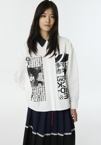 Camicia bianca con dettagli in stampa nera, tra cui un'immagine grafica e del testo. La camicia ha un colletto nero ed è abbottonata.