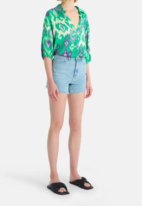 Zielona i biała bluzka w wzory z głębokim dekoltem w kształcie V, w połączeniu z jasnoniebieskimi szortami dżinsowymi i czarnymi sandałami z wiązaniem.