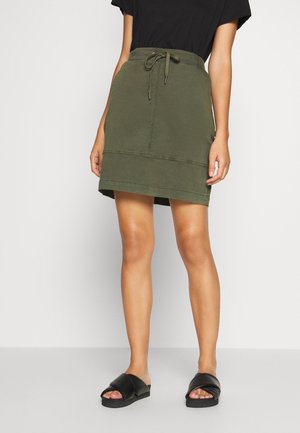 Khaki zielona spódnica z wiązanym w pasie, prosty krój i kontrastowe przeszycia na dolnej krawędzi. Nosi ją w czarnych klapkach.