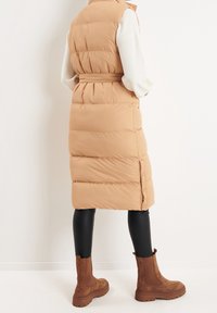Long, beige puffer vest with a belted waist. Textured, quilted design. Paired with black leggings and brown, suede ankle boots.