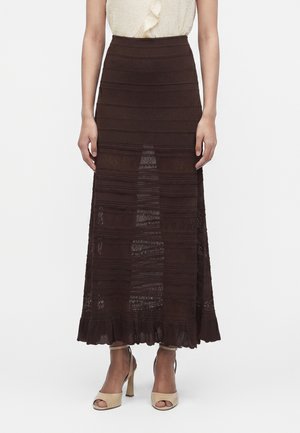 Žena nosí dlhú sukňu s vysokým pásom, tmavohnedú s čipkovaným vzorom a volánovým lemom a béžové sandále s otvorenou špičkou na bielom pozadí.