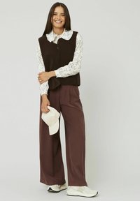 Gilet en polaire marron sur une chemise à col floral blanche, associé à un pantalon large marron et une casquette claire. Des baskets blanches complètent la tenue.