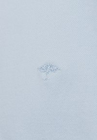 Lichtblauwe katoenen stof met een klein geborduurd boomlogo in een iets donkerdere tint, centraal op het kledingstuk geplaatst.