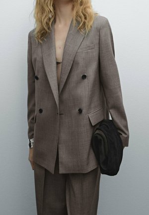 Femme portant un costume marron oversize à double boutonnage tenant une veste noire matelassée pliée sur un fond clair uni.