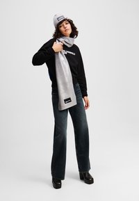 Silver metallic scarf draped over a black sweatshirt with text. Model wears wide-leg dark jeans and black platform boots, with a grey beanie.