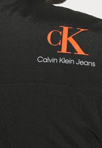 Czarna kurtka o gładkiej teksturze, z wyraźnym pomarańczowym logo "cK" oraz napisem "Calvin Klein Jeans" w białym kolorze w pobliżu ramienia.