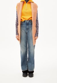 Pletená oranžová vesta s kapsami přes květinovou košili s dlouhým rukávem, doplněná vysoce pasovými bledými modrými džíny a černou obuví.