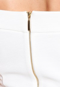 Bílá žebrovaná tkanina se středovým zlatým zipem a malou nálepkou poblíž jezdce, ukazující texturu a detail.