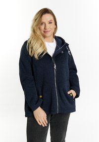 Mėlyna zip-up stiliuje striukė su tekstūruotu paviršiumi, reguliuojama kapučiu ir dviem priekinėmis kišenėmis, turinčiomis geltonus mygtukus. Dėvima virš baltos marškinėlių.