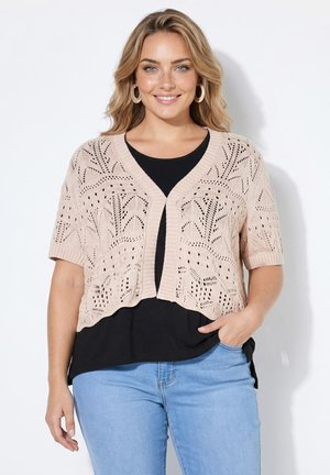 Kvinde med blondt hår bærer en beige kortærmet strikket cardigan over en sort top og lyseblå jeans, smilende med den ene hånd i lommen.