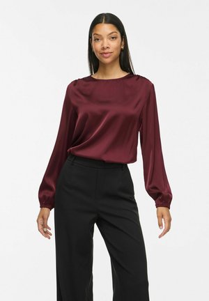 Vrouw draagt een bordeauxrode satijnen blouse met lange mouwen en zwarte, hoog gesneden wijde broekspijpen, staand tegen een effen achtergrond.